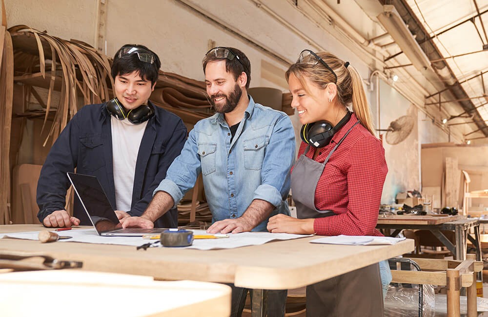 Drei Handwerker in Schutzbrillen und Gehörschutz stehen an einem Tisch mit Bauplänen und Laptop in einer Werkstatt mit Holz und Werkzeugen.