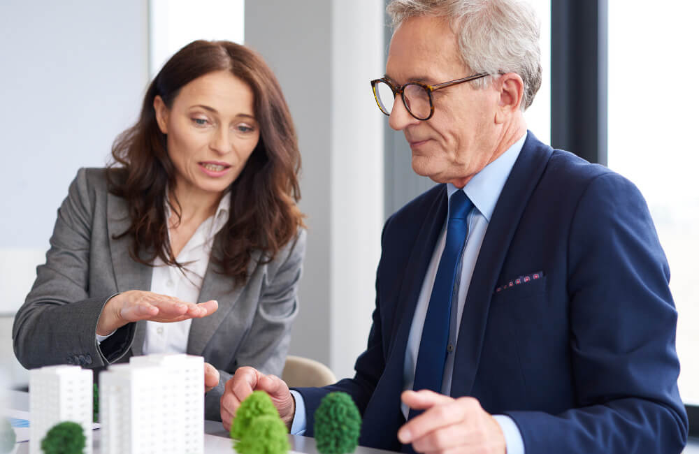 Zwei Personen in Businesskleidung sitzen an einem Tisch mit Architekturmodell und Miniaturbäumen, diskutieren offenbar Immobilien.