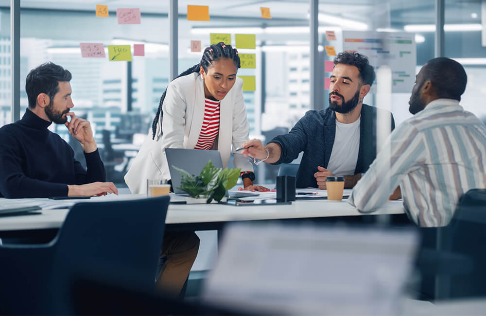 Vier Personen sitzen an einem Tisch in einem modernen Büro, eine Person steht und zeigt auf einen Laptop, bunte Haftnotizen kleben an der Glaswand im Hintergrund.